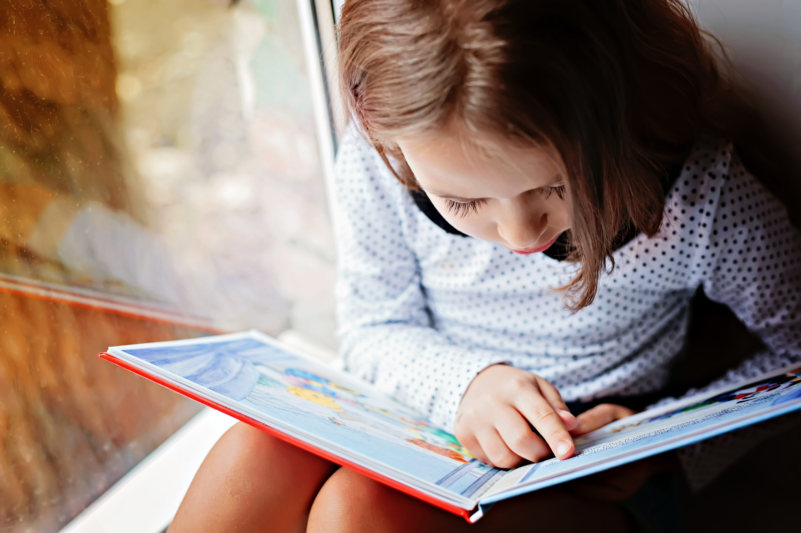 Toddler,Girl,With,Book,Near,The,Window